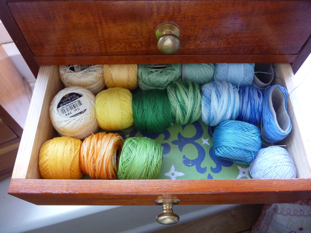 A miniature wooden chest of drawers with one drawer open displaying spools of thread in yellows, greens and blues