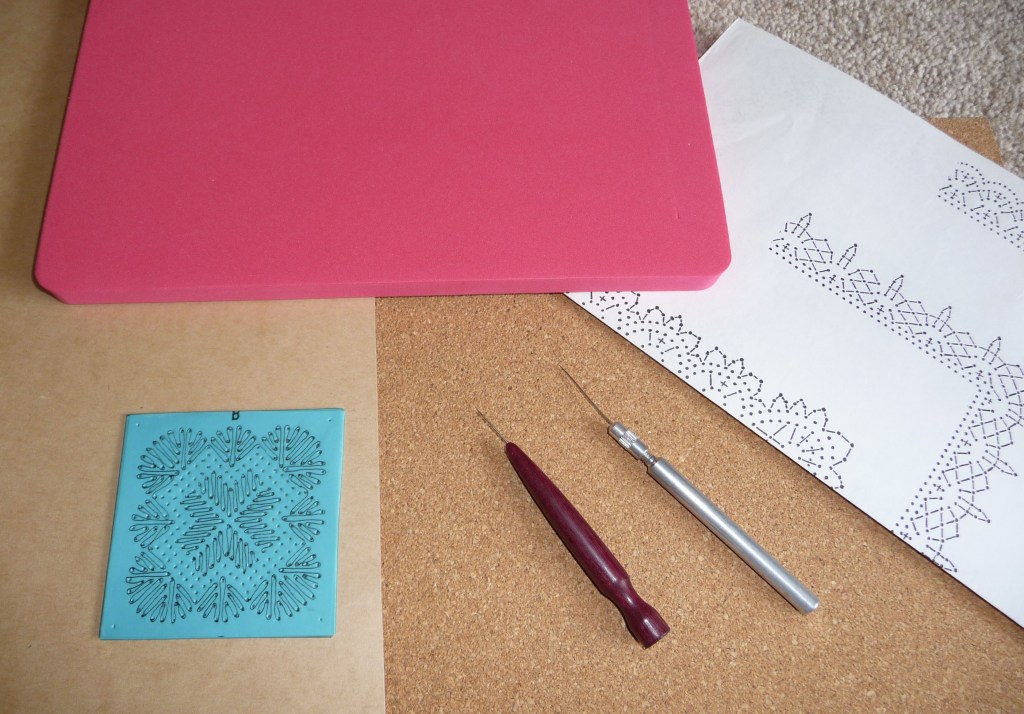 A selection of equipment used for pricking out a lace pattern including a cork pricking board, a memory foam pricking board, a metal pricking tool and a wooden handled tool. 