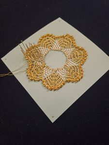 A small circular piece of lace using white and gold threads with a border of gold beads